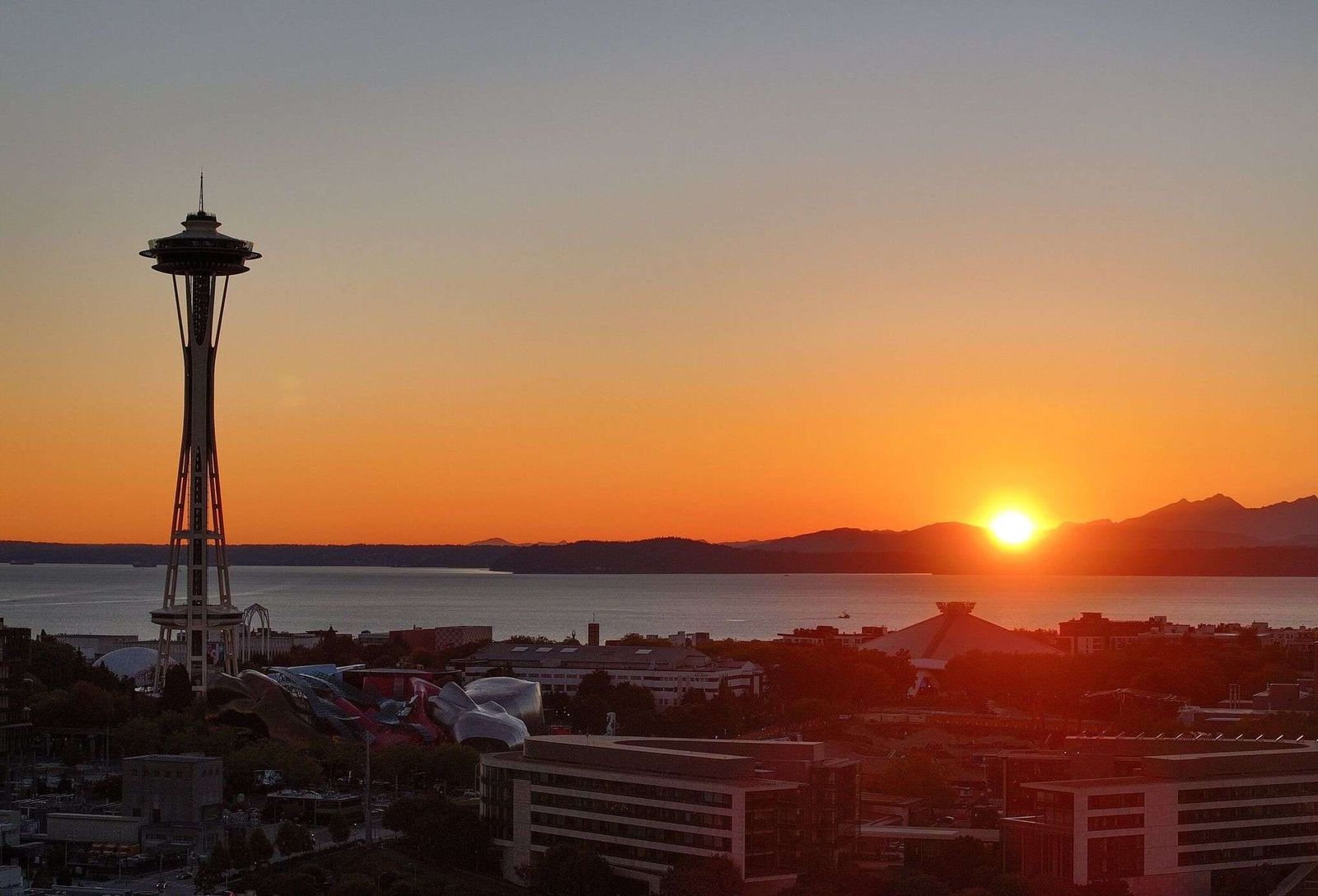 The Space Needle silhouetted against an orange sky as the sun sets over Elliott Bay and the Olympic Mountains, with Seattle Center below